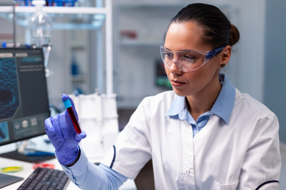 Pesquisadora em laboratório vestindo jaleco, luvas e óculos de proteção analisa um tubo de ensaio com amostra de sangue, representando estudos e diagnósticos de doenças autoimunes.