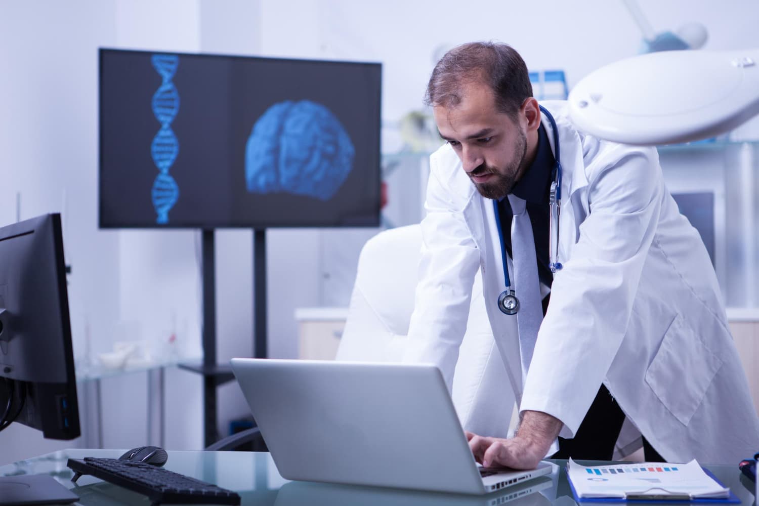 O que e neurologia: médico neurologista em seu consultório olhando o notebook. Na parece, um monitor com o mapeamento de um cérebro