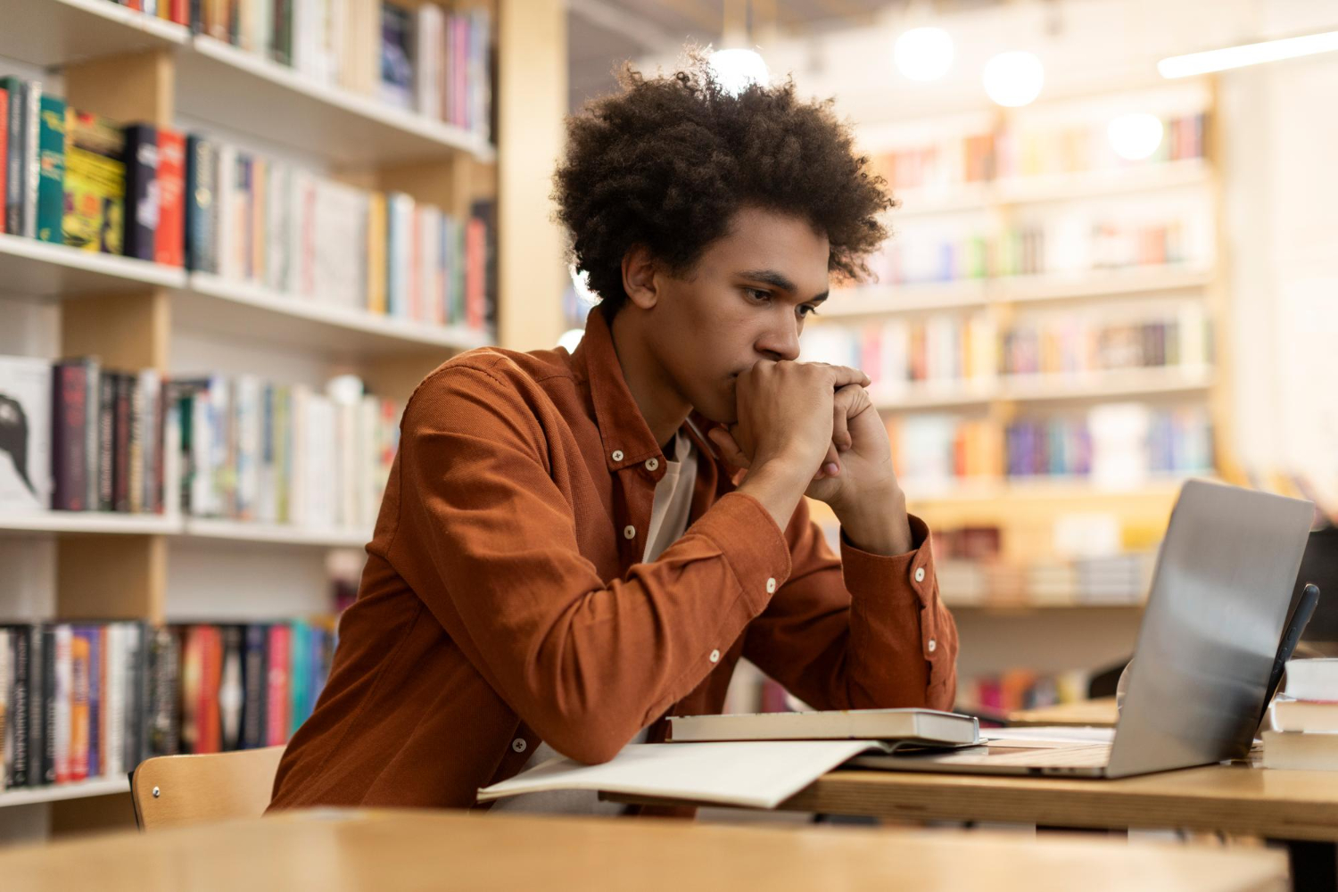 Produtividade acadêmica: Jovem estudante na biblioteca de uma faculdade, concentrado à frente do seu notebook e de um livro aberto.