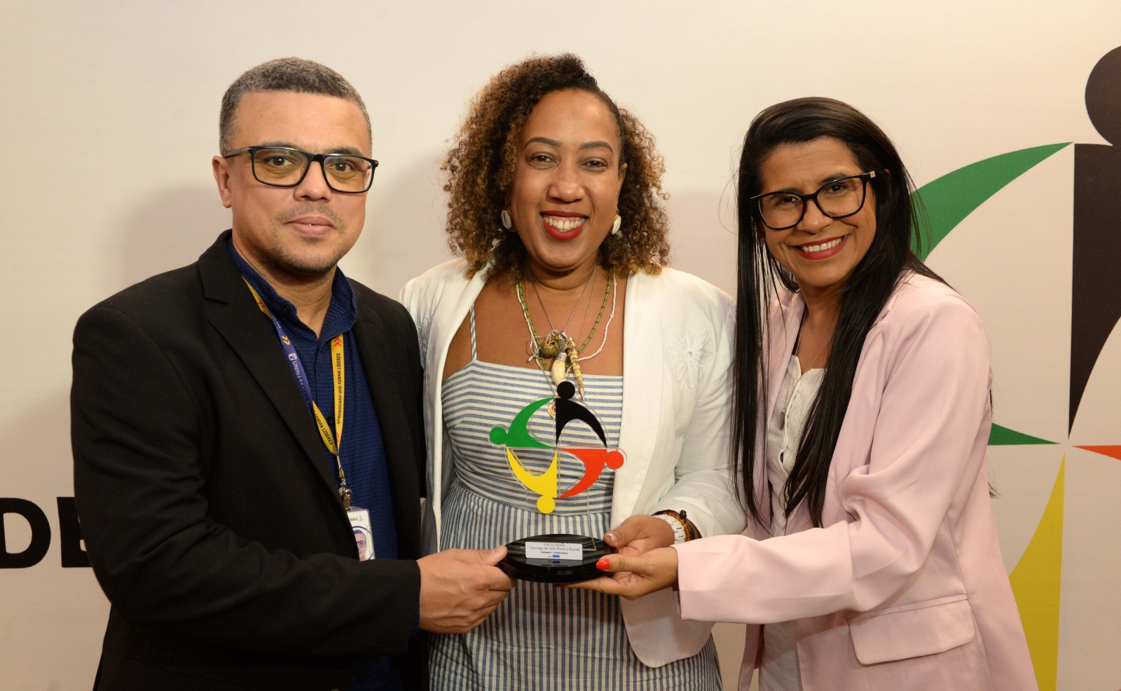 Três pessoas posam com um troféu durante cerimônia de reconhecimento em evento sobre diversidade étnico-racial.
