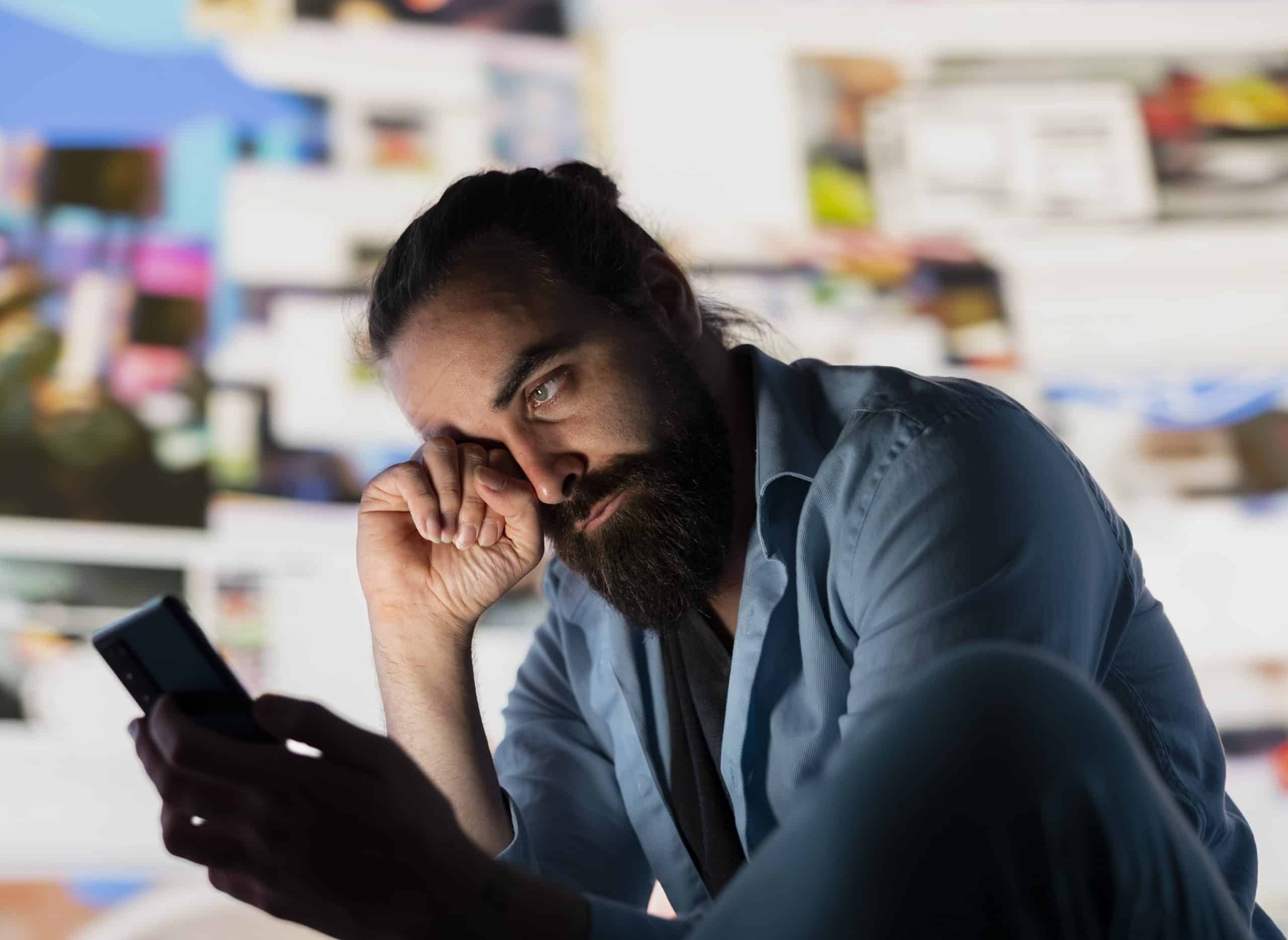 Homem sentado olhando para o celular com expressão de cansaço e preocupação, representando os impactos do excesso de tempo de tela na saúde mental e no bem-estar.